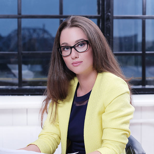 Frau mit langen Haaren und Brille, die in einer gelben Jacke am Schreibtisch sitzt und in die Kamera schaut.